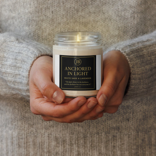 “Hands gently holding a white glass candle labeled ‘Anchored in Light’ by JB, featuring a black and gold design. The candle scent is White Sage & Lavender, with the scripture John 1:5 printed on the label. A warm, soft light glows from the flame, creating a cozy, faith-inspired atmosphere.”