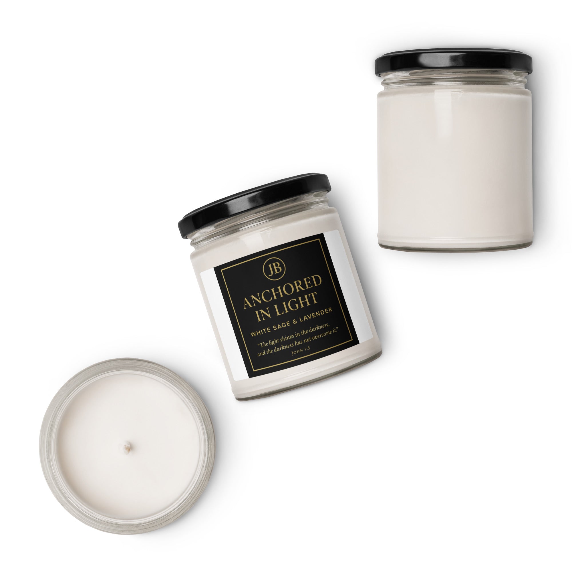 “Three ‘Anchored in Light’ white glass candles with black lids arranged on a white background. One candle is lying on its side, showing the JB black and gold label with scripture John 1:5, while the other two are upright and open to display the wax inside. Minimal, clean, luxury product display.”